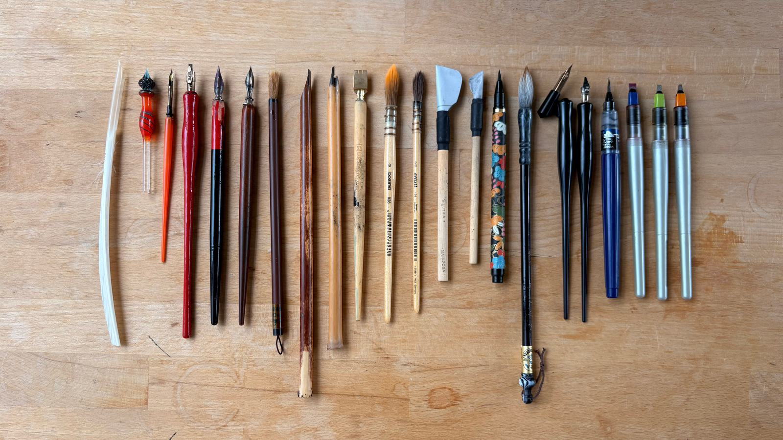 Pinceles, plumas y cálamos alineados sobre una mesa de madera.